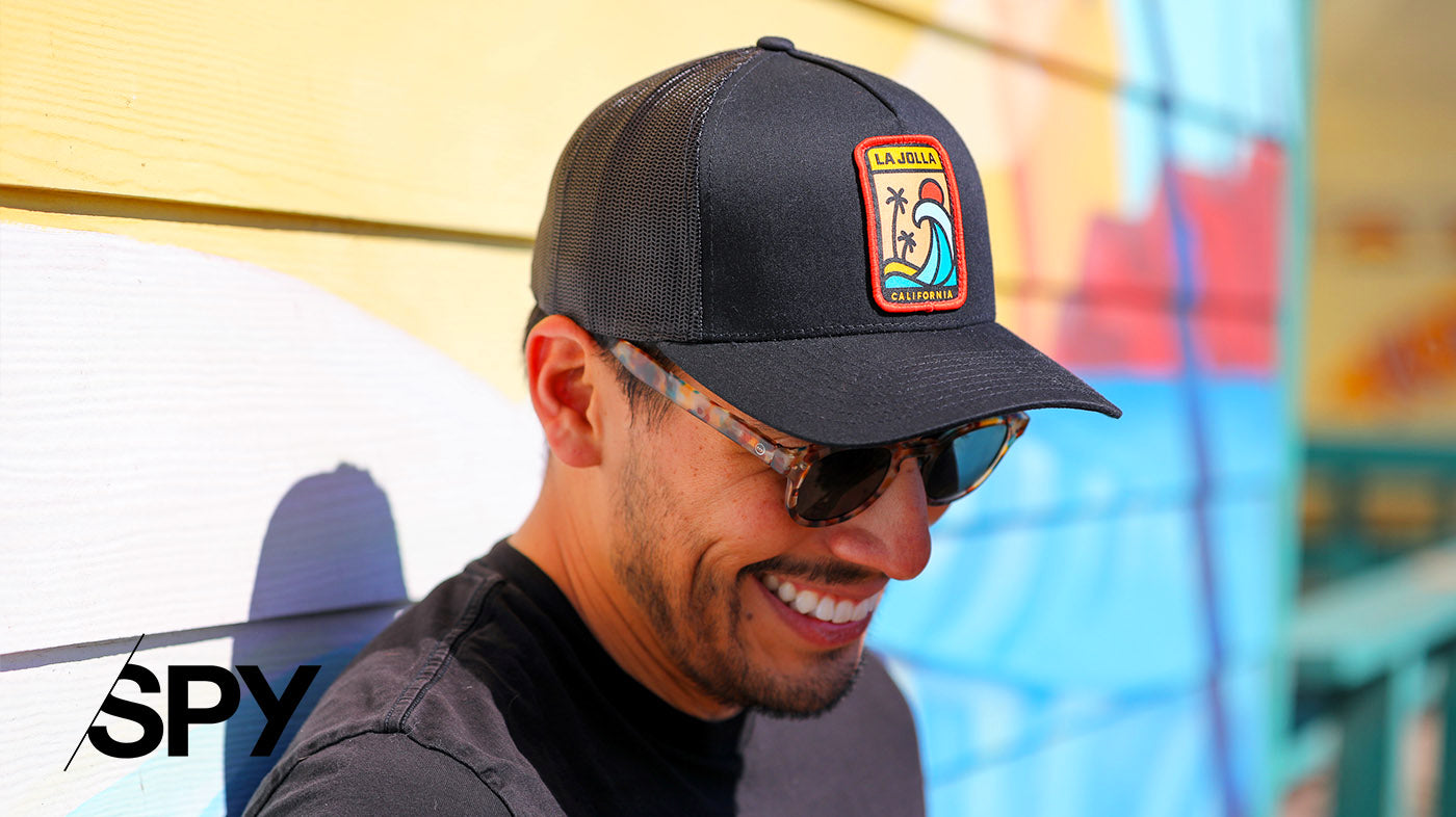 Photo of a man wearing a hat and smiling, leaning against a brightly painted wall