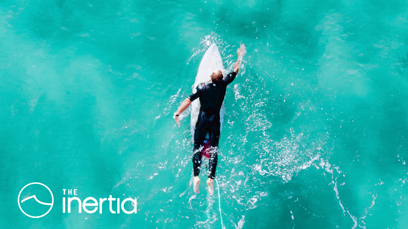 Aerial photo of a surfer in a wetsuit paddling through bright blue water on a white surfboard